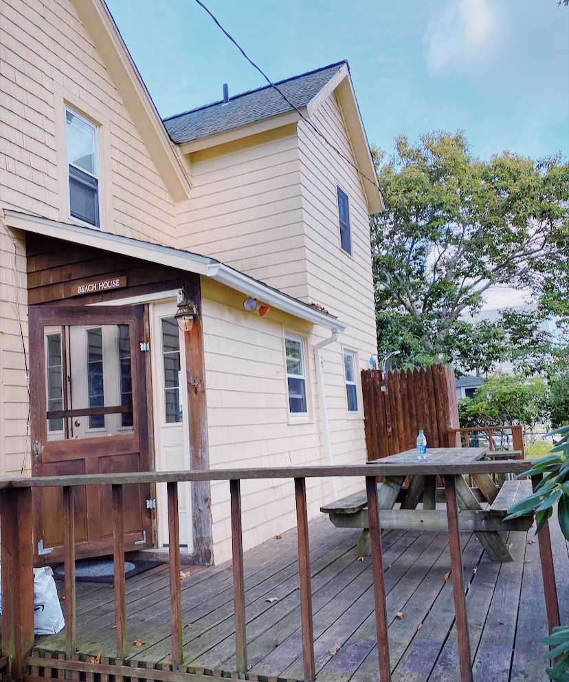 This photo depicts a yellow house with a back porch and picnic table beneath lush green trees