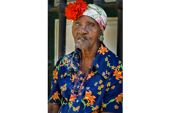 Older woman in bright floral top, with a flower on her hat, smoking a cigar