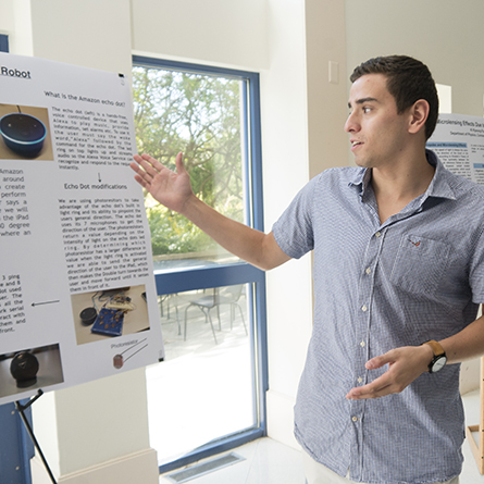 George Tilneac '18 describes his research building an autonomous double robot while pointing at a poster that explains the project.