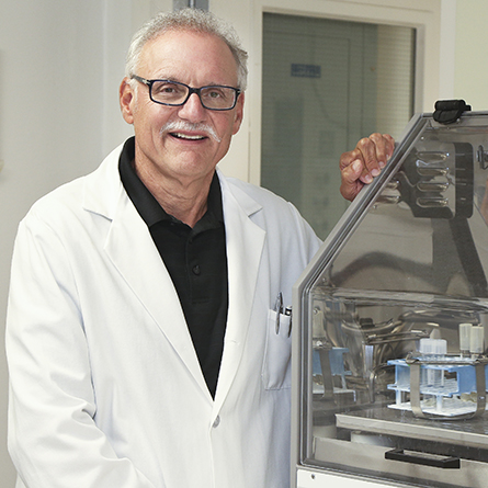 Professor Bruce Branchini, wearing a lab coat, stands next to a large piece of lab equipment. 