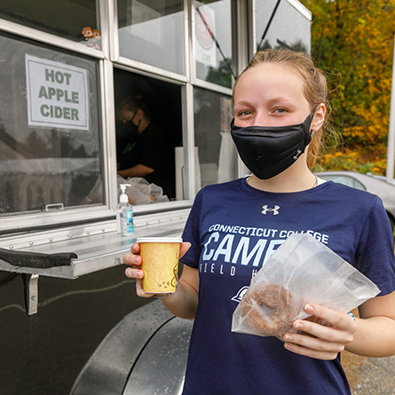 A student holds a donut and hot apple cider outside of a food truck.