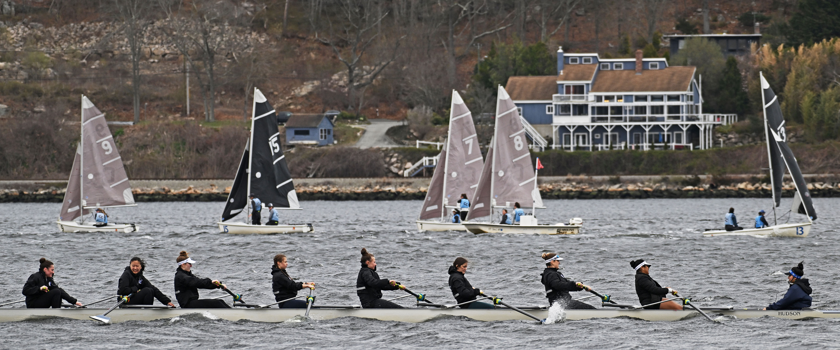 Far shot of crew team rowing in front of CC sailboats