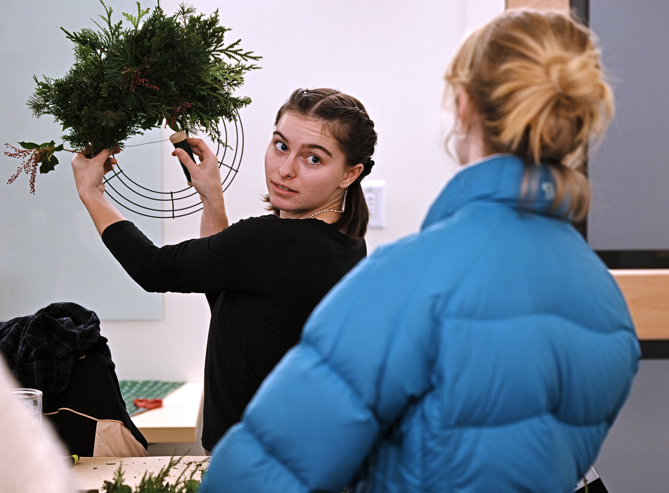 A student holds up a wreath in progress to show another student during a wreath-making workshop.