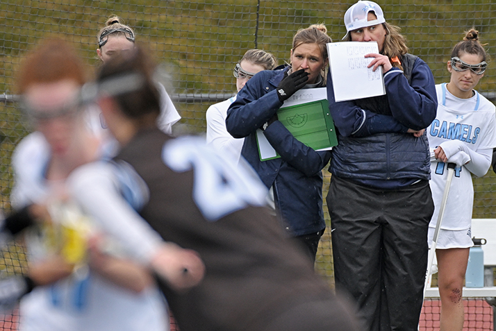 two lacrosse coaches talk to each other on the sideline as game action takes place in the foreground