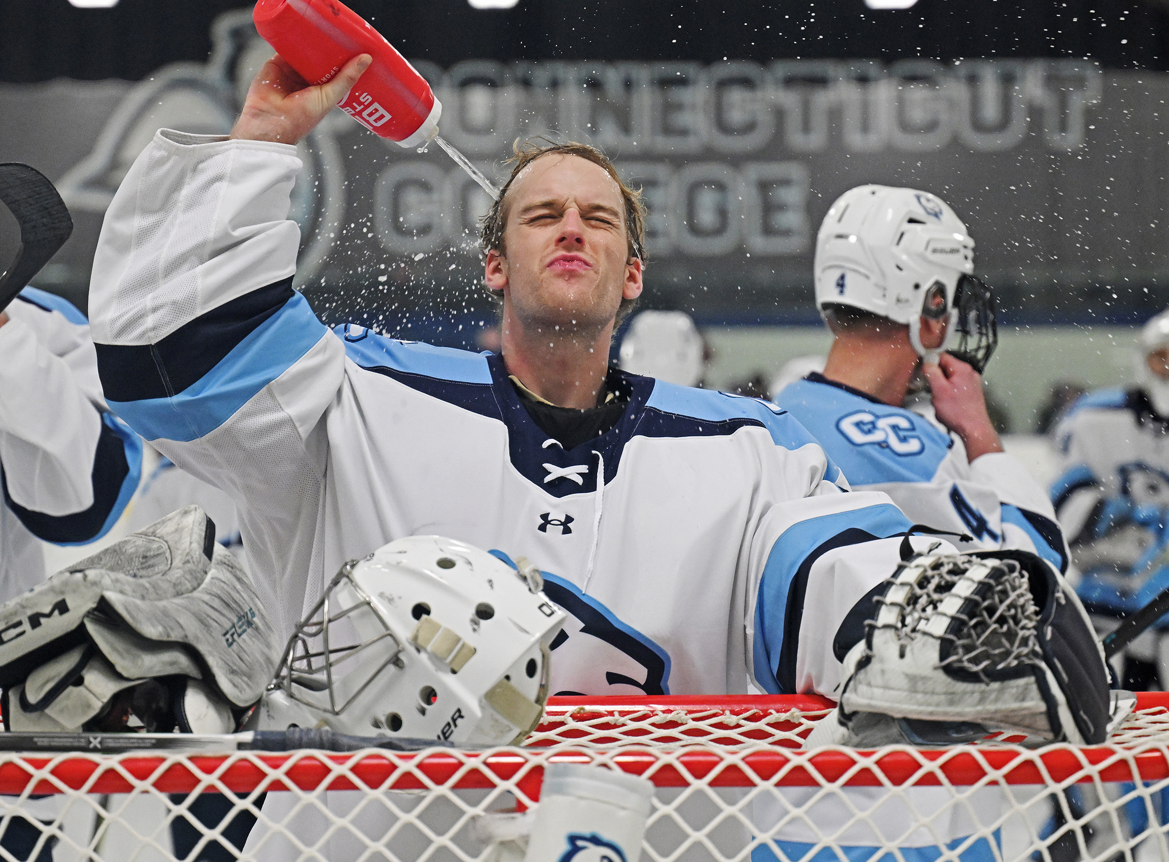 A hockey goalie sprays himself with his water bottle standing over the goal.