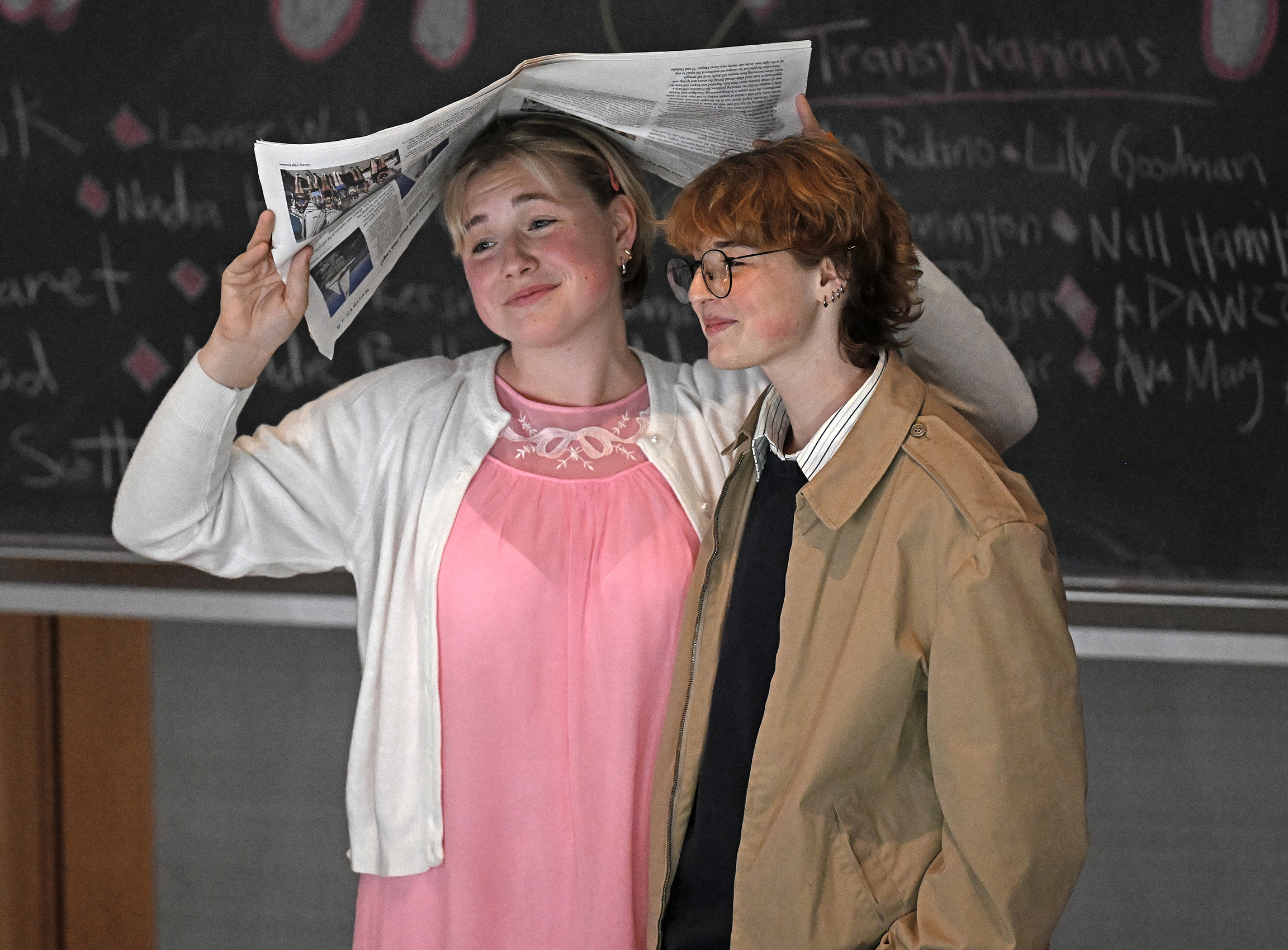Two student actors, one in a pink dress and cardigan, and one in khaki jacket, shelter under a newspaper during a performance of The Rocky Horror Picture Show.