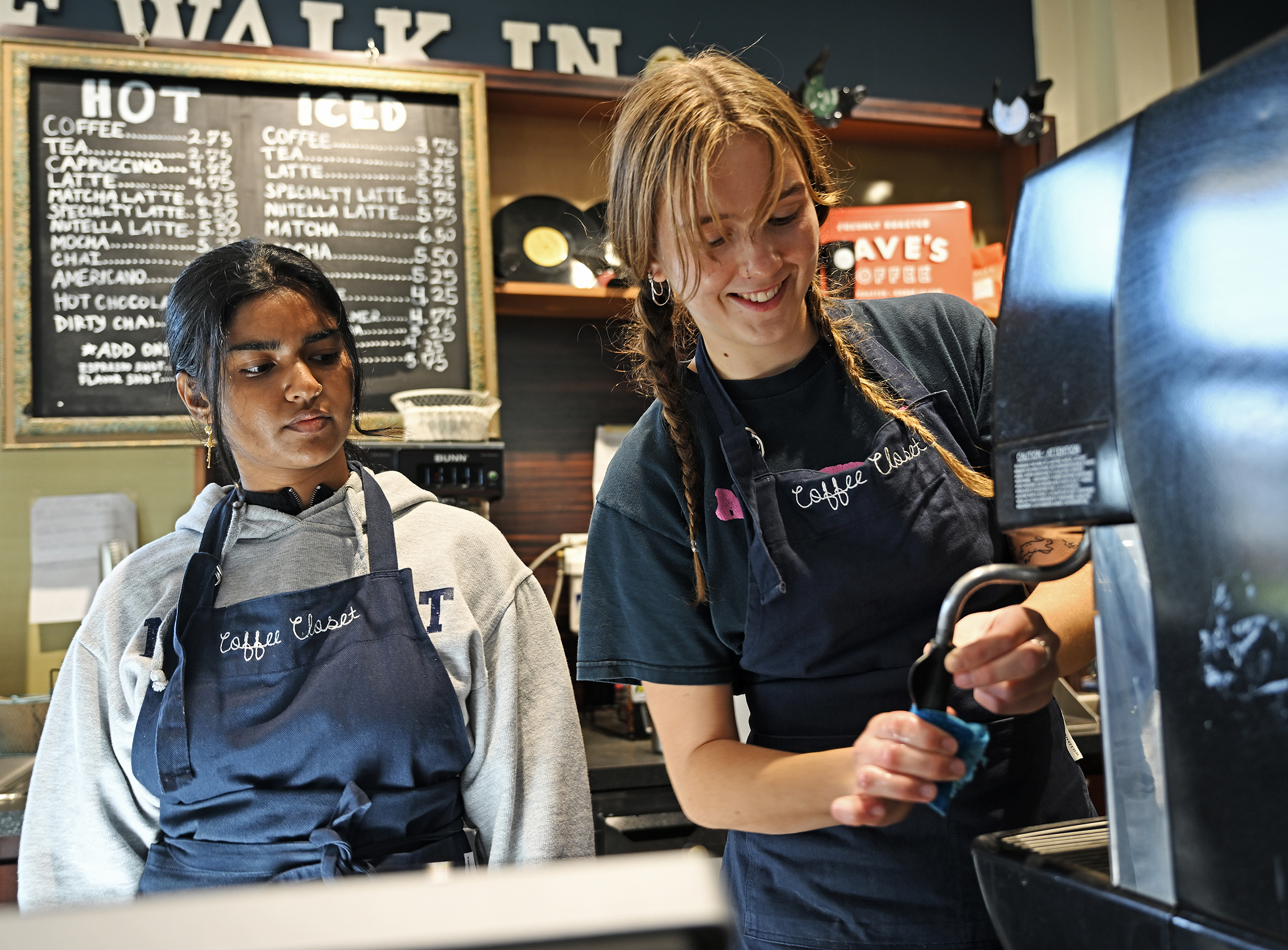 One student watches as another works at an espresso machine in a coffee shop.