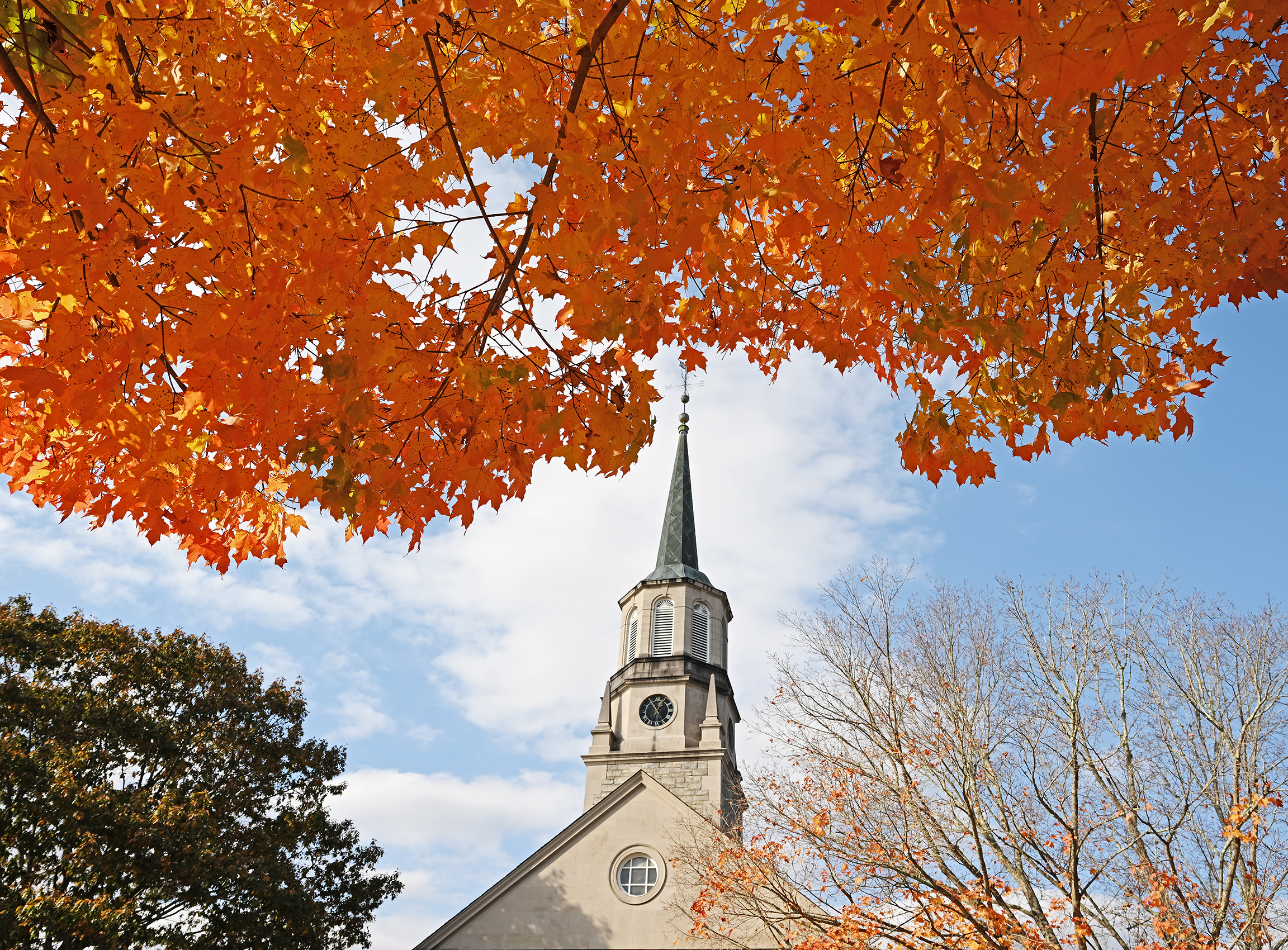 Bright red/orange maple leaves frame a chapel steeple on a clear fall day.