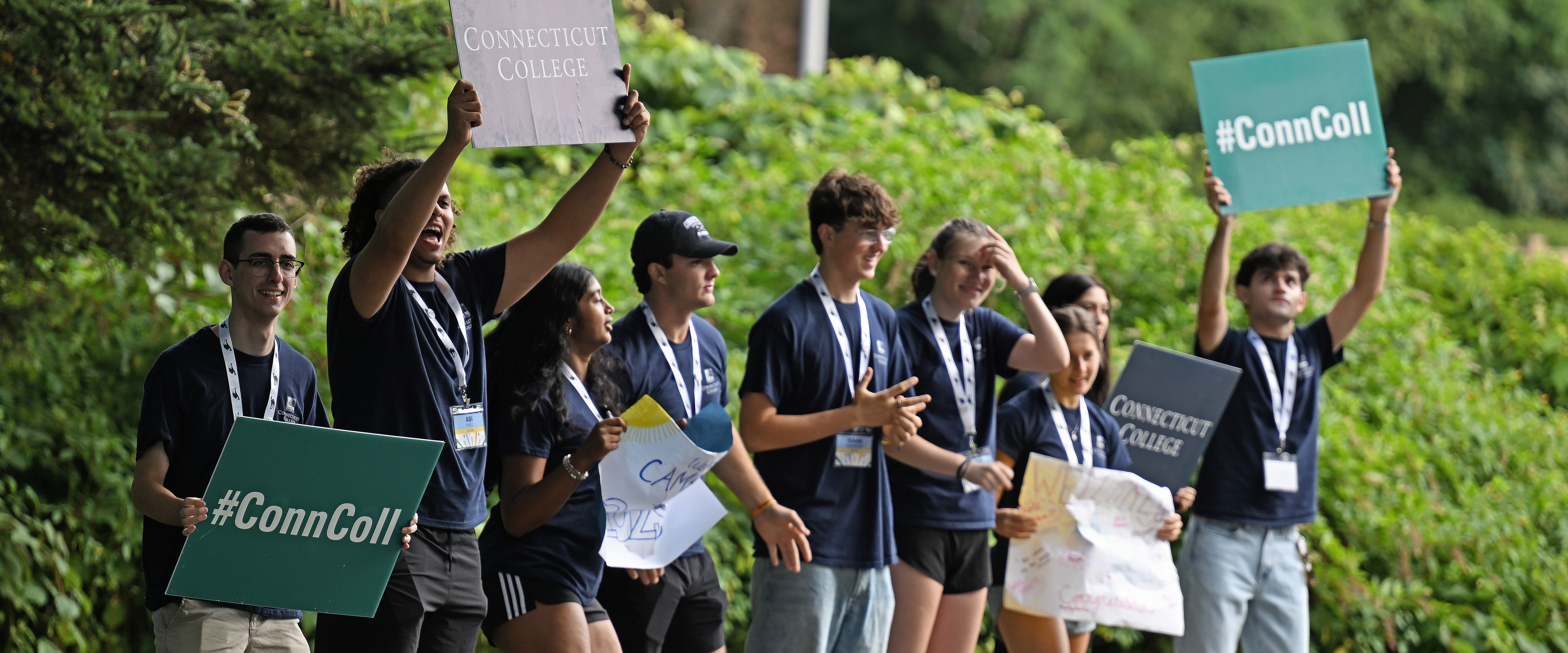 A group of students holding signs cheer for new students arriving on campus.