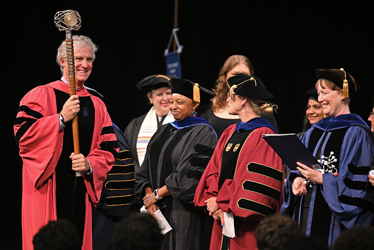A college professor in academic regalia carries a ceremonial mace during a convocation ceremony.