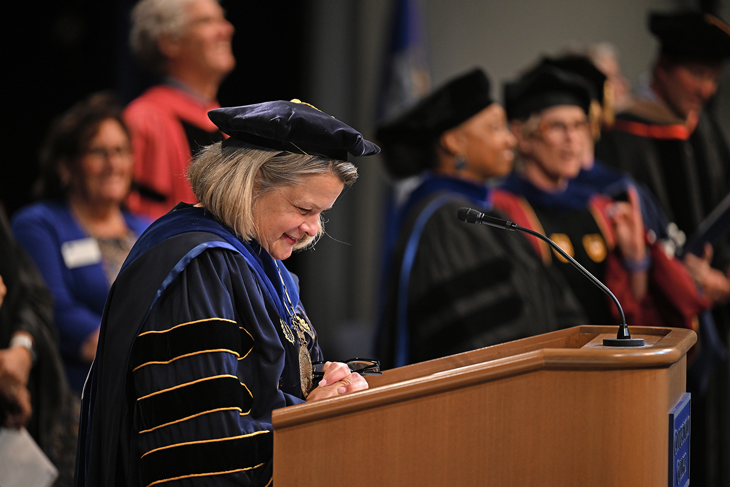 A college president in academic regalia laughs while speaking at a podium
