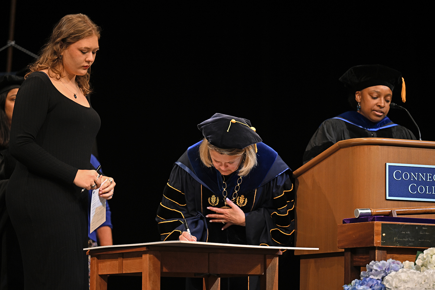 A college president in full academic regalia bends over to sign a document on stage during a convocation ceremony.