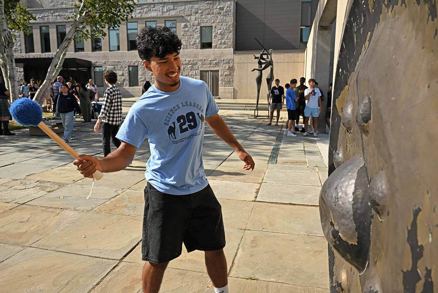 A student in a blue t-shirt rings a giant gong hanging on the wall of an outdoor courtyard.