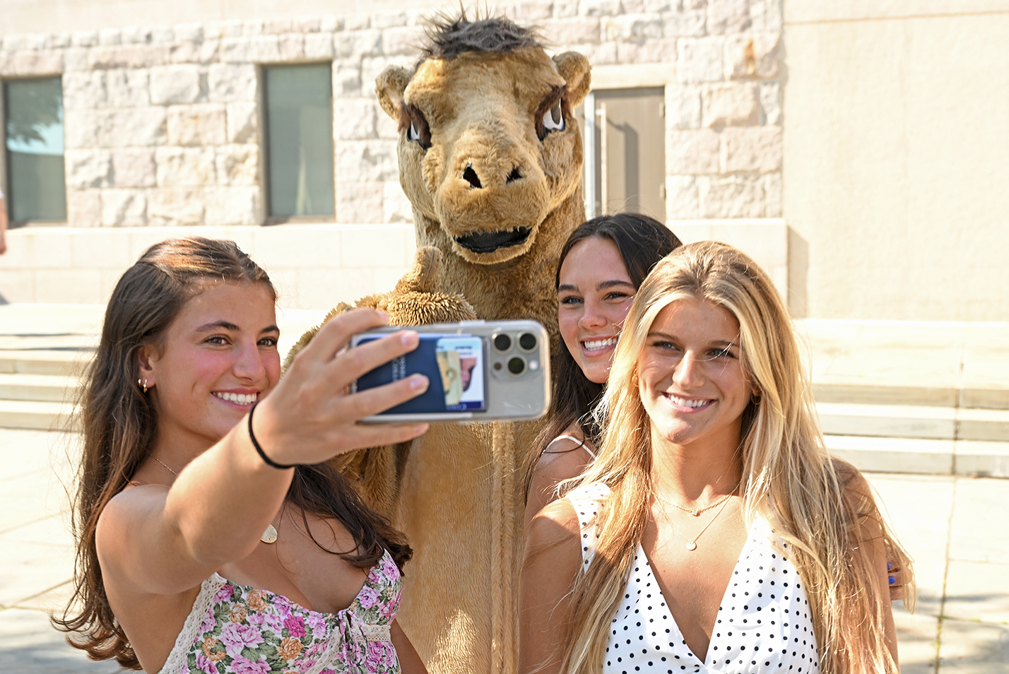 Three female students in sun dresses take a selfie photo with a college mascot camel.