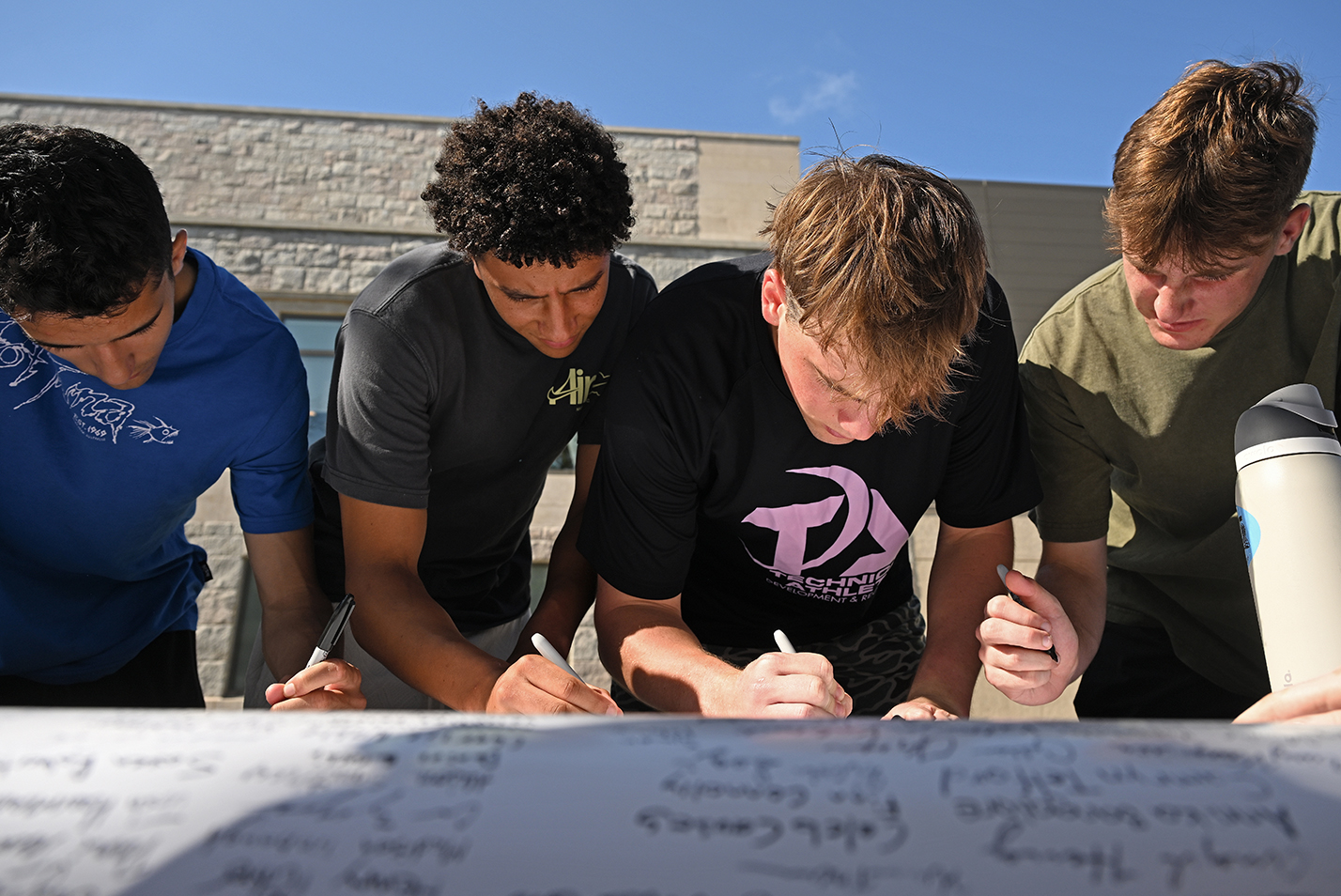 Four male college student lean over to sign a post-sized document on a table in an outdoor courtyard.