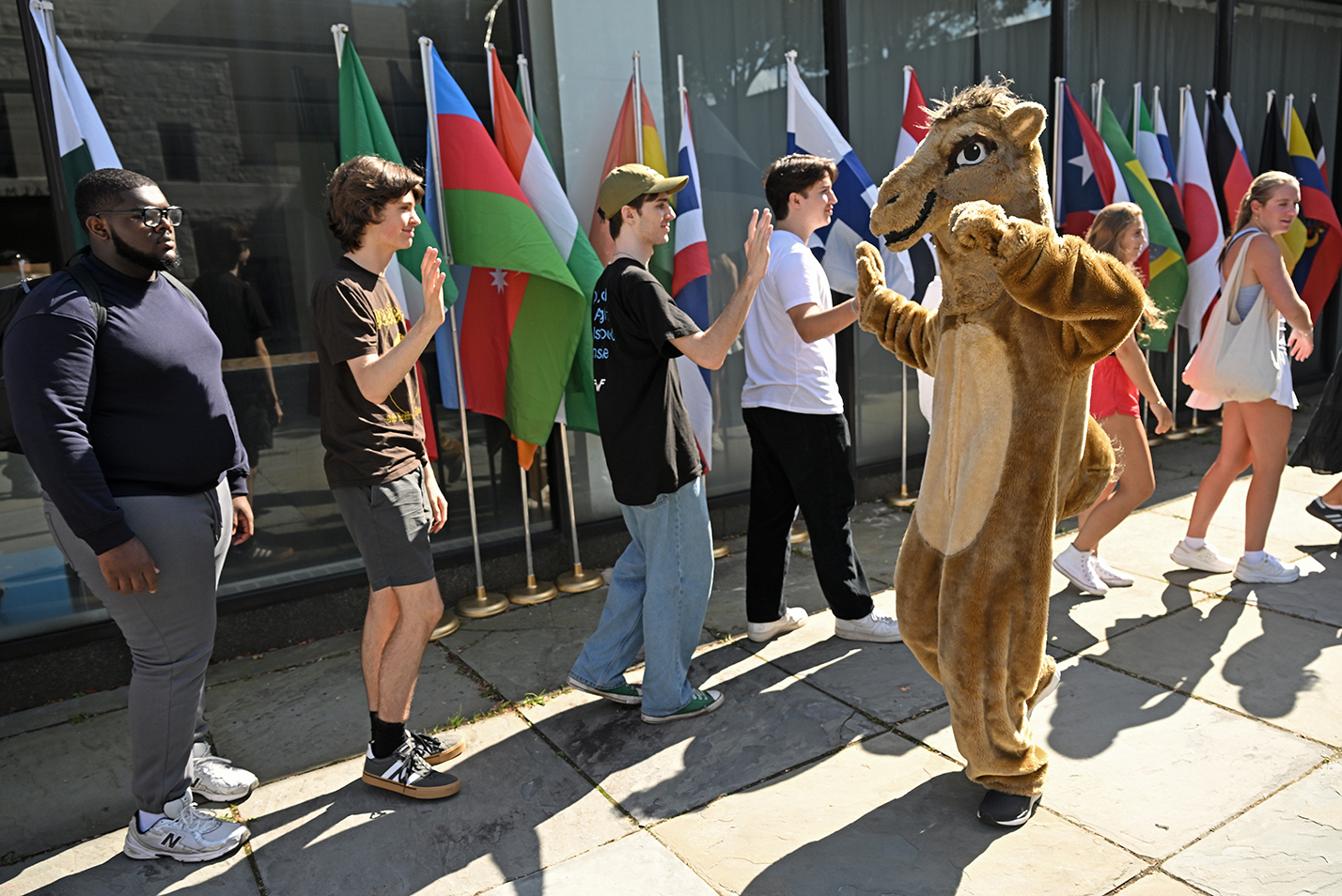 A college mascot camel high-fives students standing in a line in front of a display of international flags.