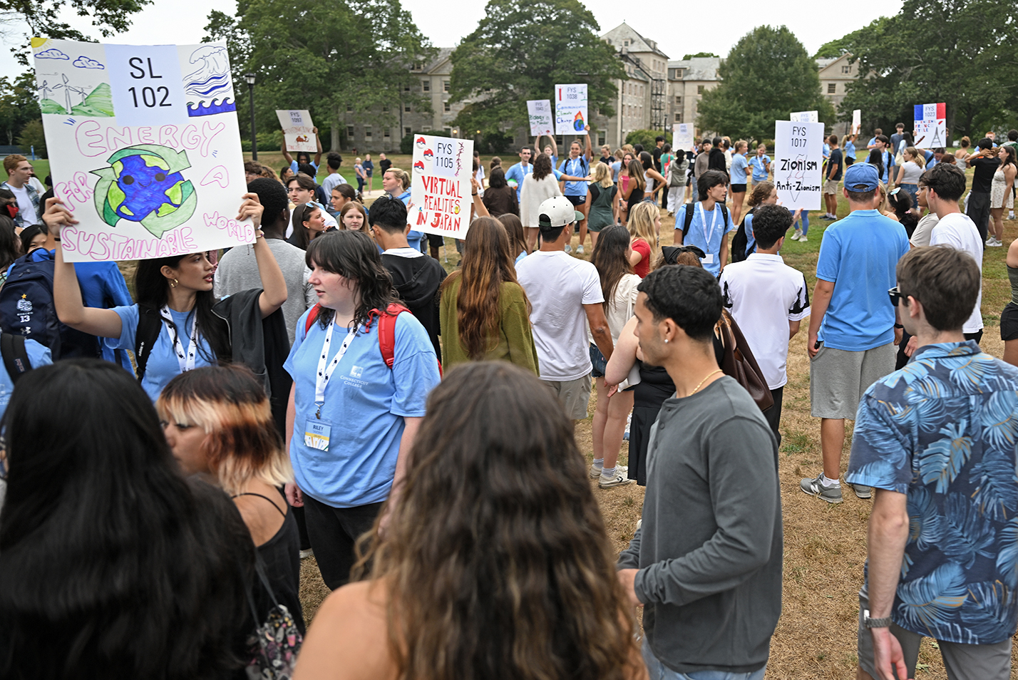 Student advisers hold signs to guide first year students to their orientation groups on a college green.