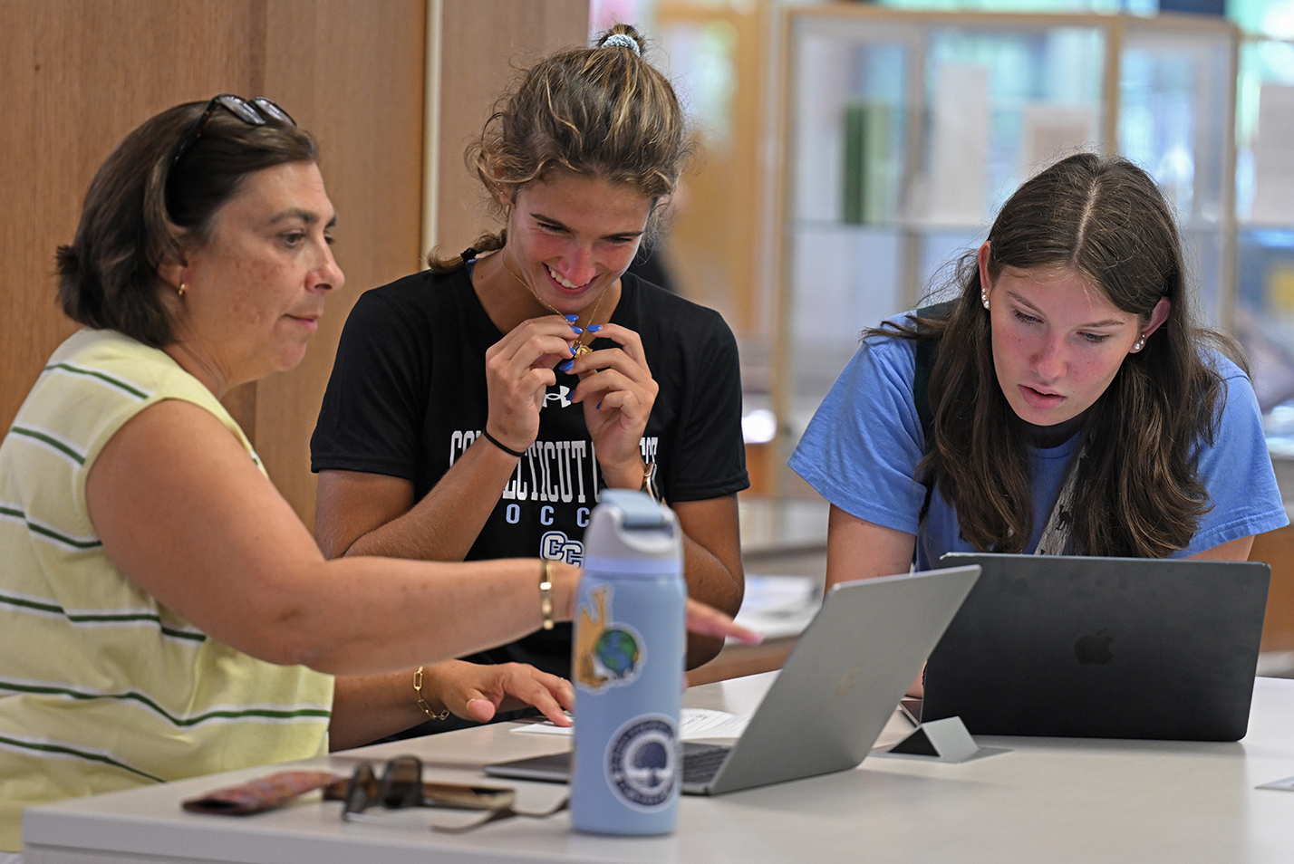 A college student smiles while getting help registering for courses on a laptop computer in a library setting.