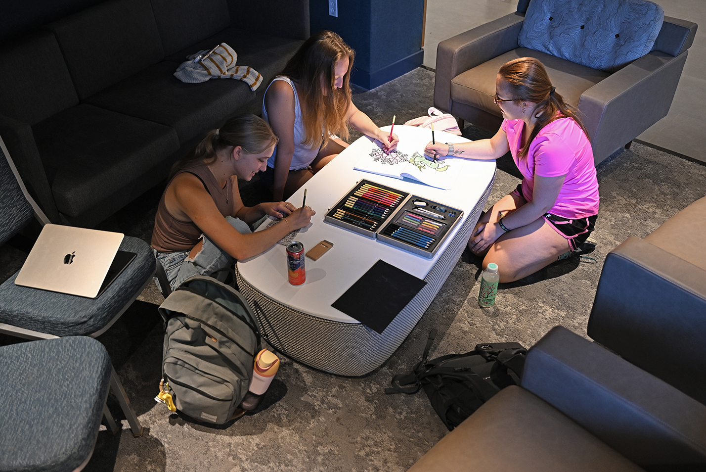 three students work in coloring books sitting on the floor around a coffee table in a lounge area.