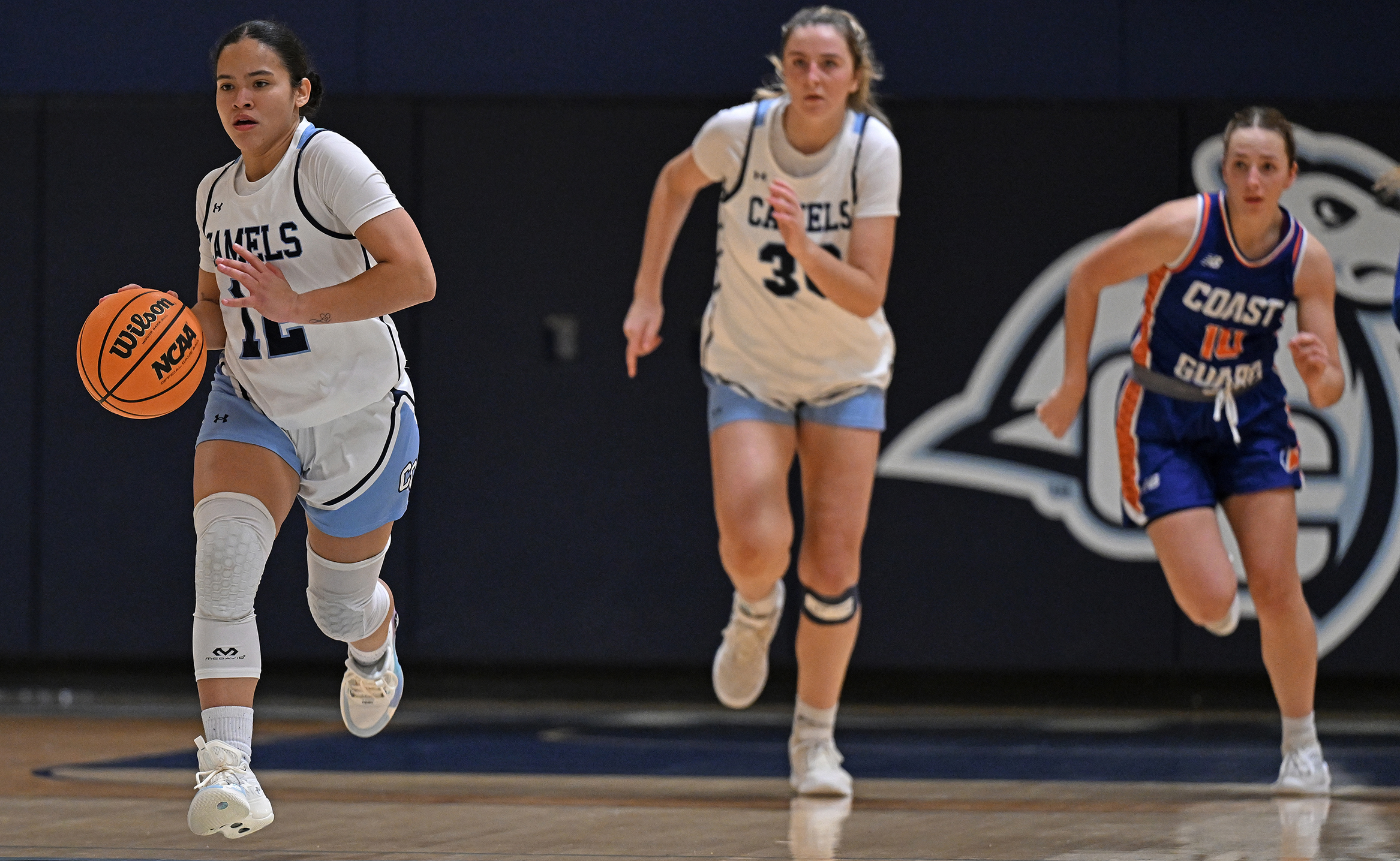 A woman basketball player dribbles the ball upcourt followed by a teammate and an opponent.