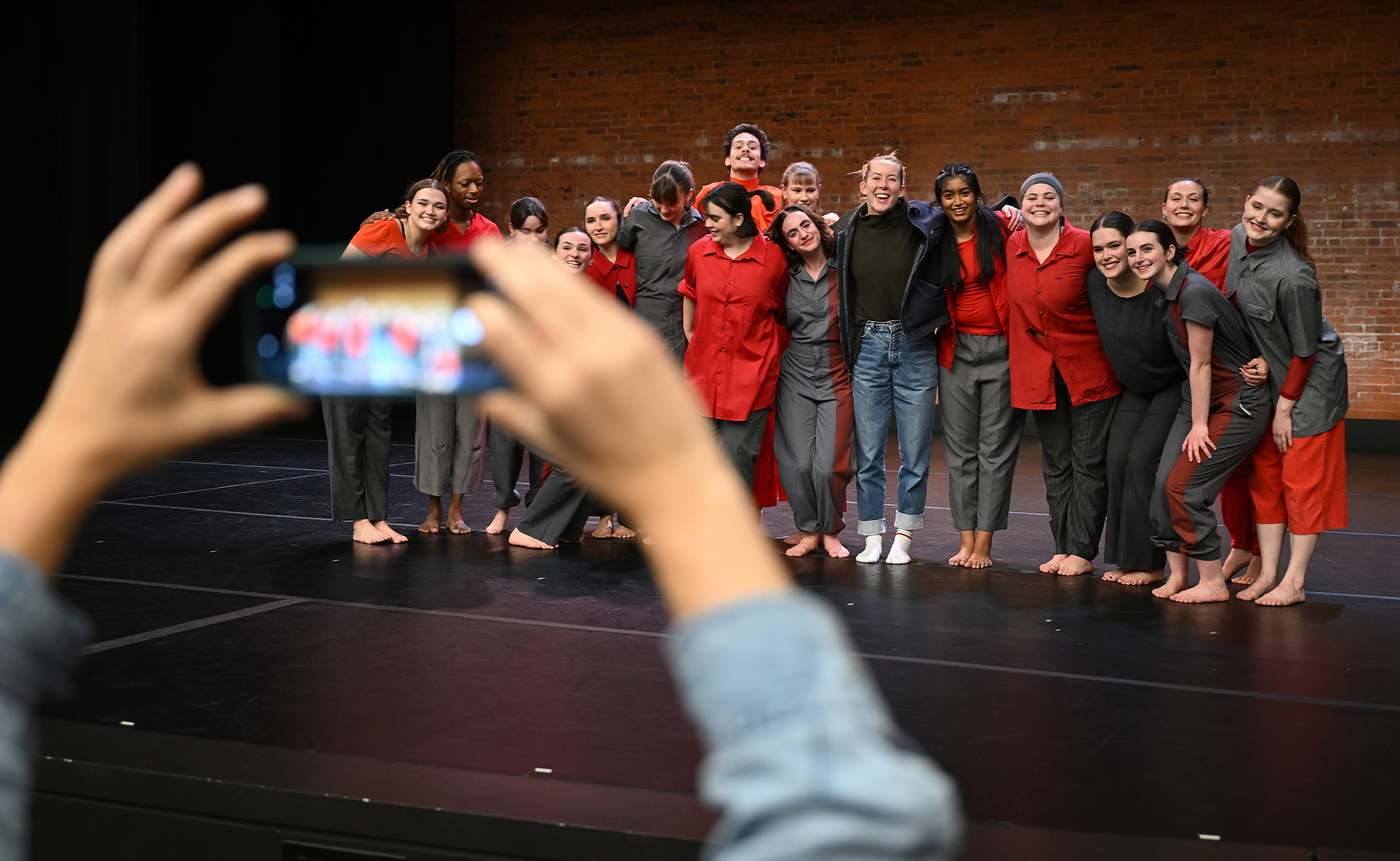 Dancers gather for a group photo on stage following a performance framed by two arms holding a smart phone overhead of the photographer.