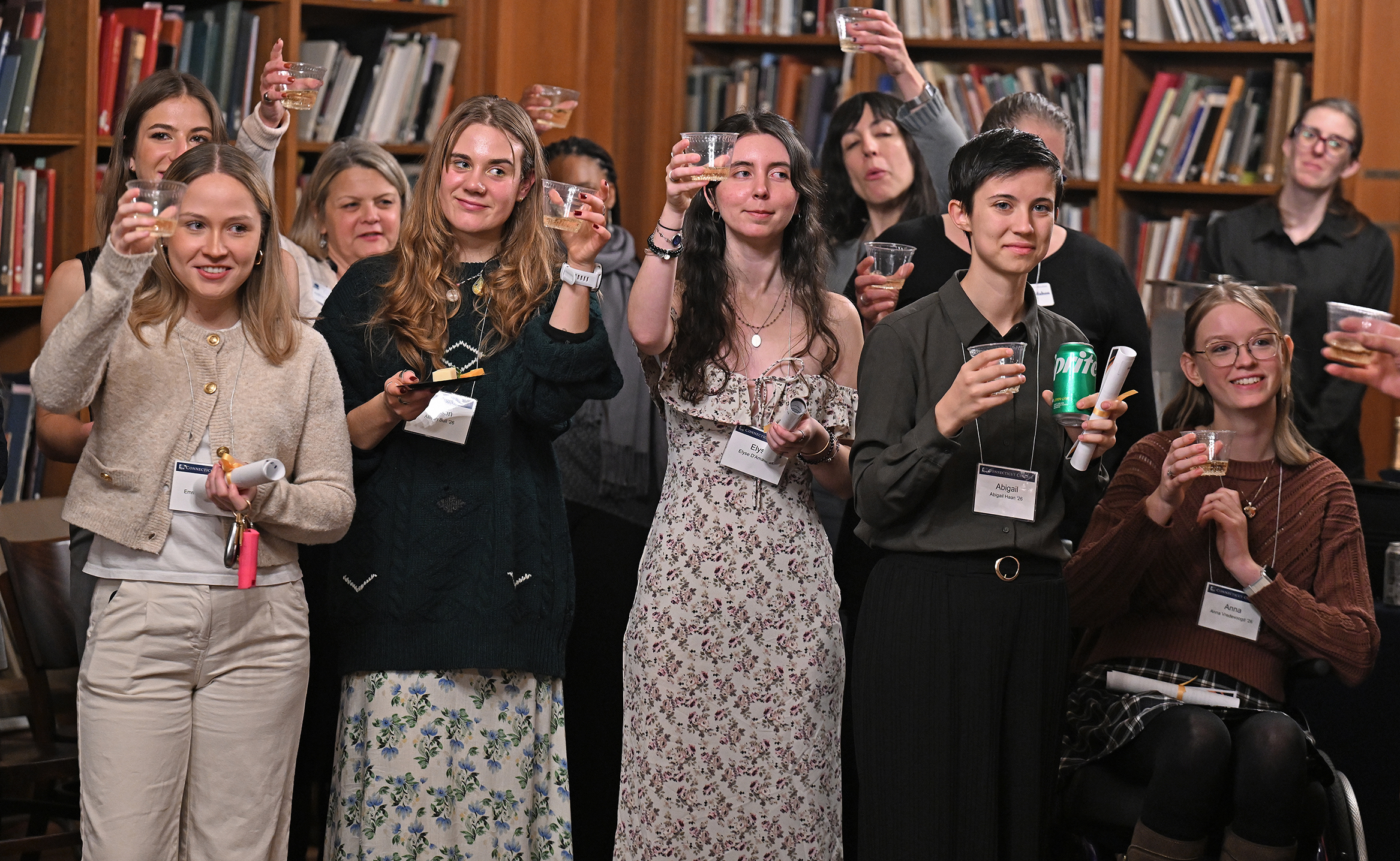 College students in semi-formal attire raise their glasses for a toast during an early graduation celebration in a formal library space.
