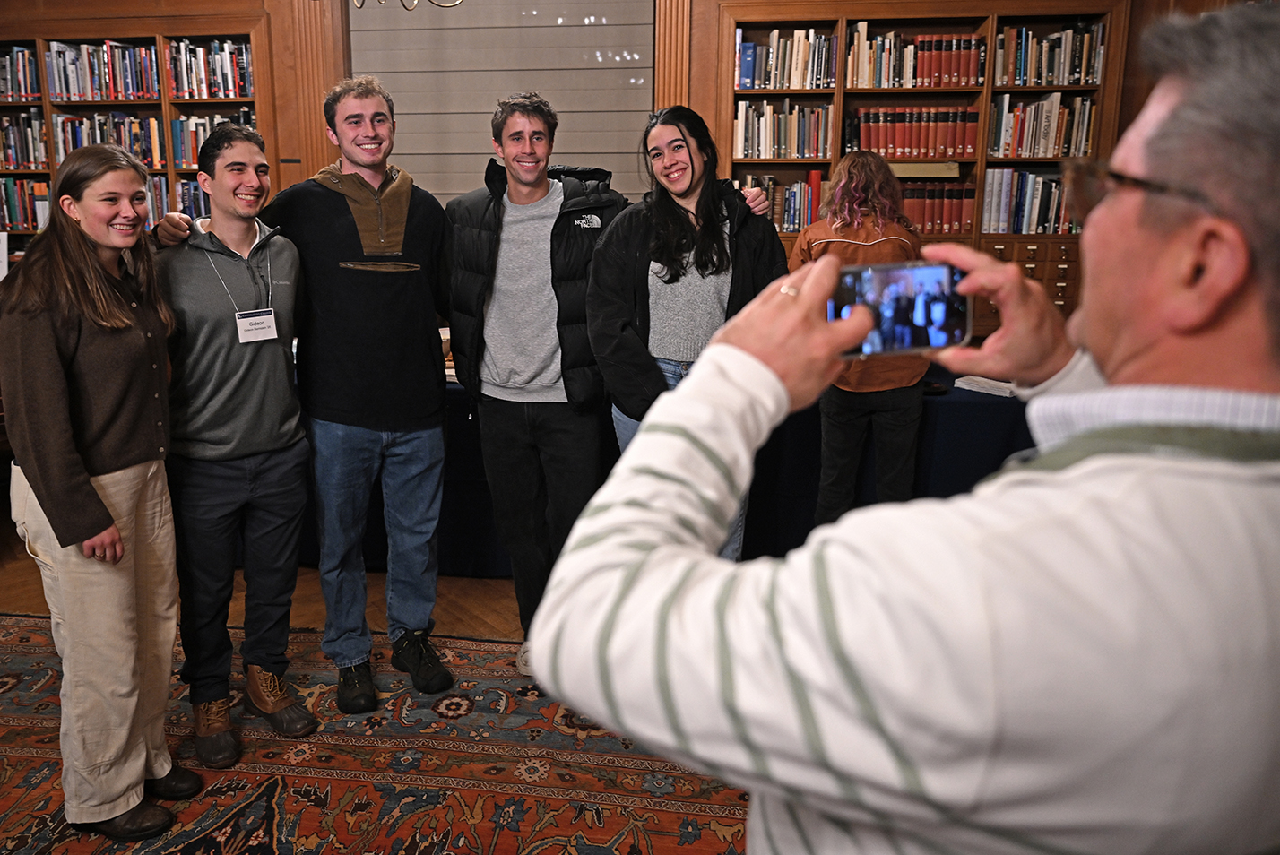 A group of early-graduating college seniors gather to take a group photo during a celebration event in a formal library setting.
