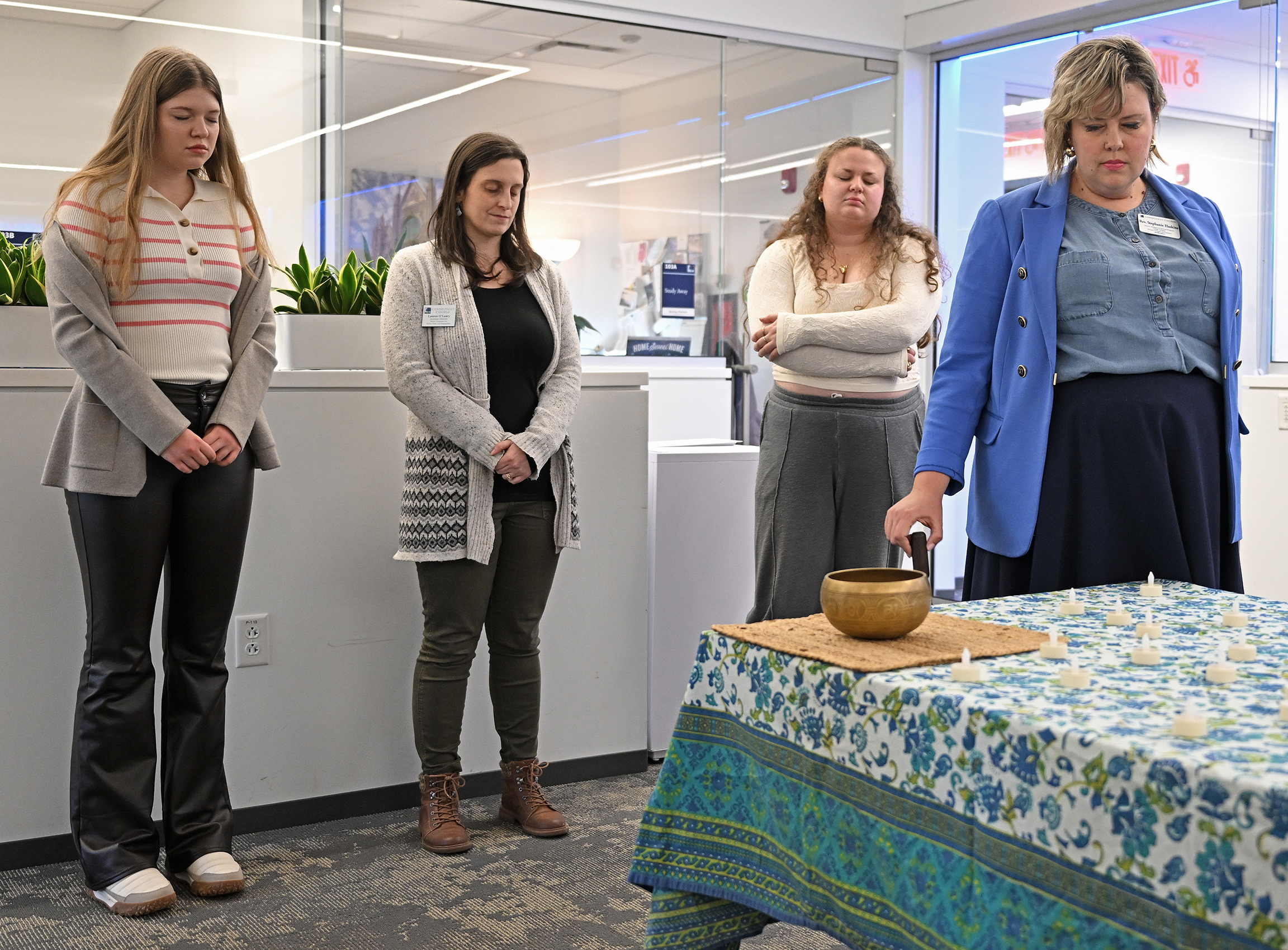 A minister rings a Tibetan singing bowl as others bow their heads in prayer during a spiritual gathering in a college common space.