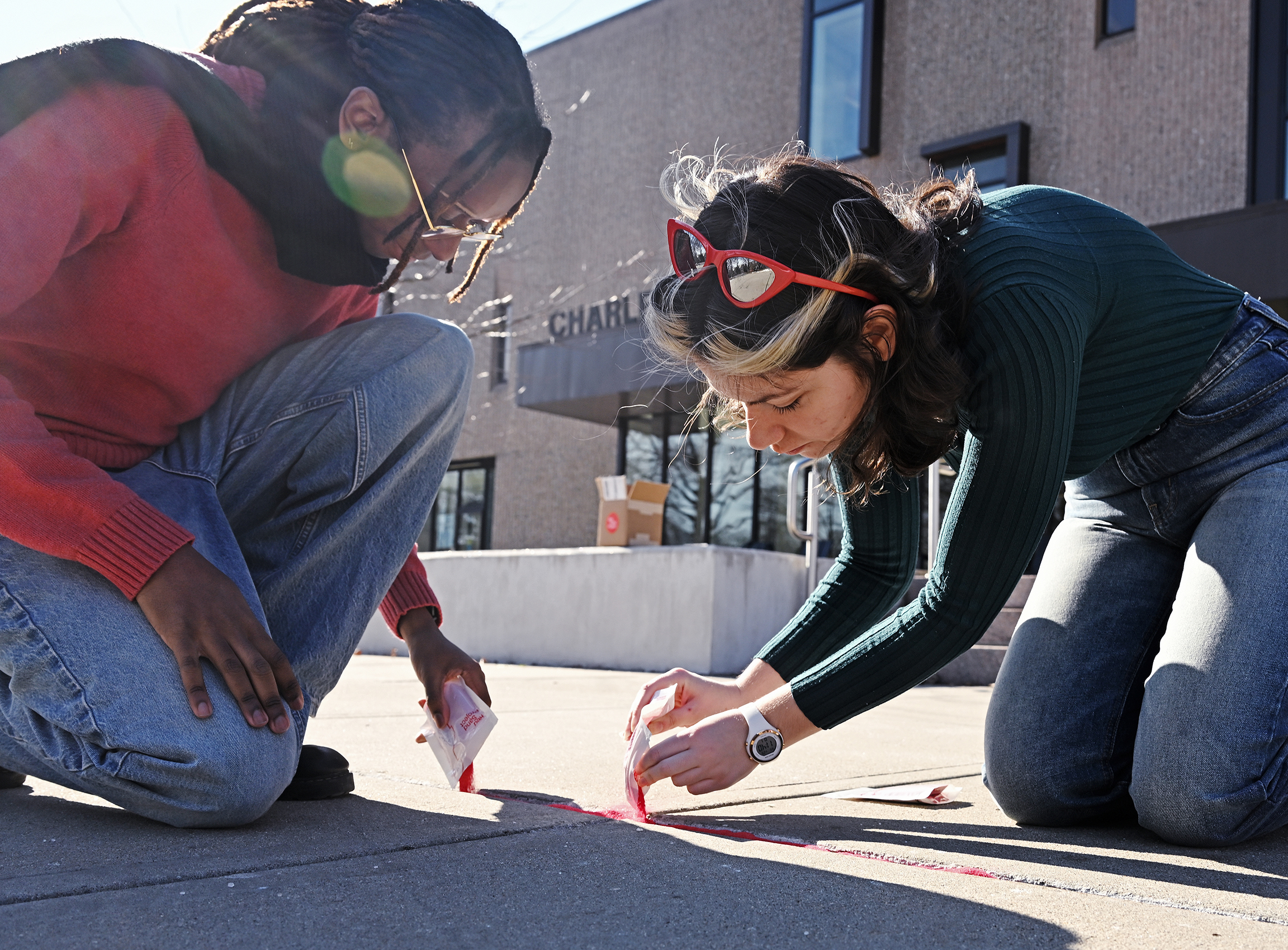 Two students kneel on the grounds and pour read sand into the seams in the sidewalk.