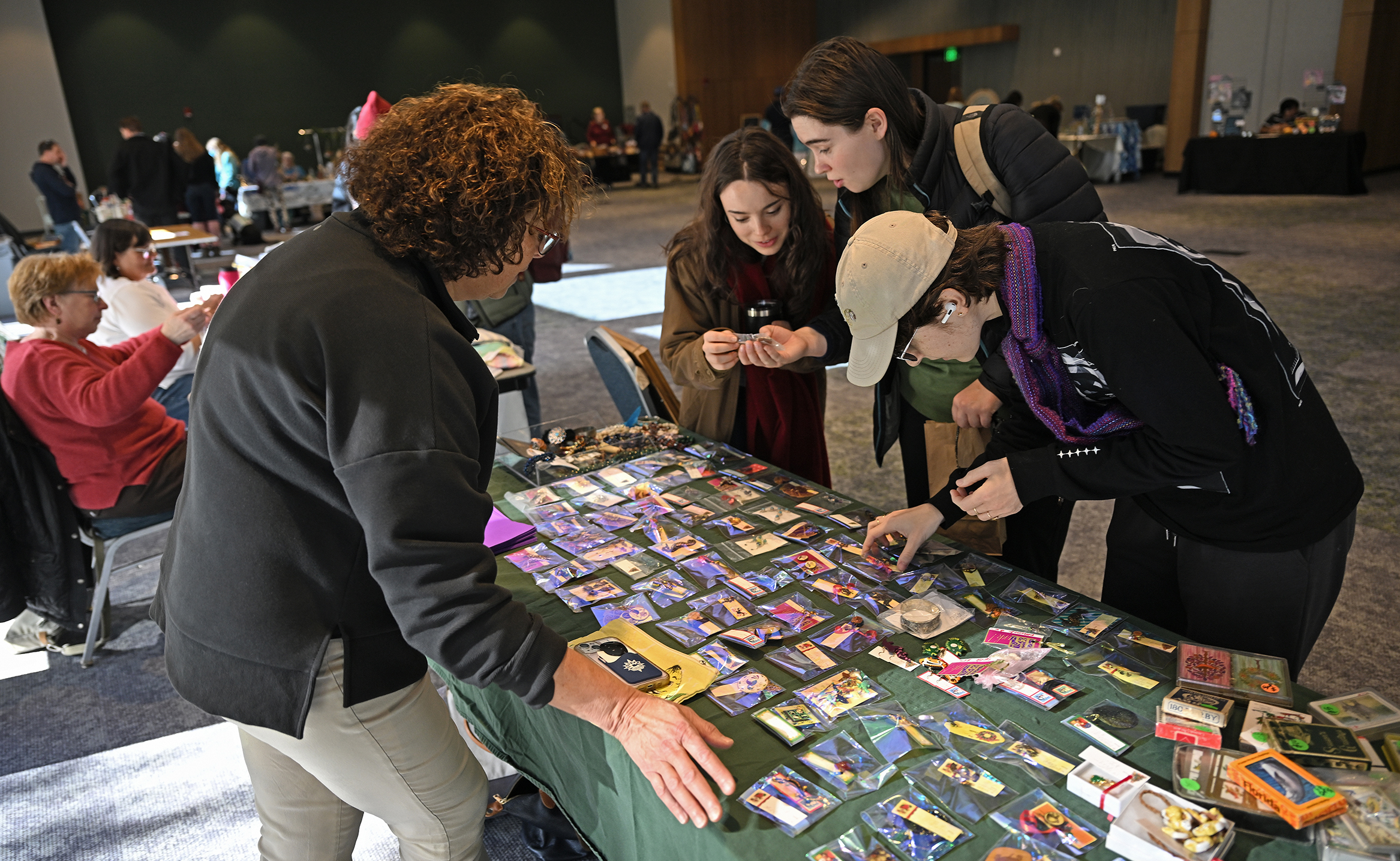 Three students look at craft items for sale at a craft fair table in an event room on a college campus.