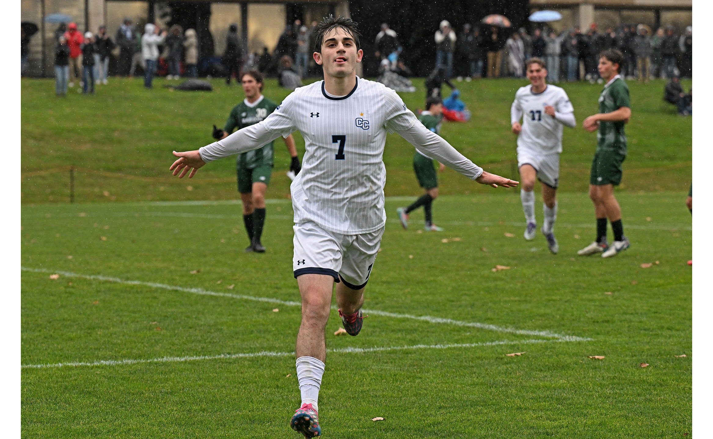 A college soccer player runs toward the sideline with outstretched arms to celebrate a goal.