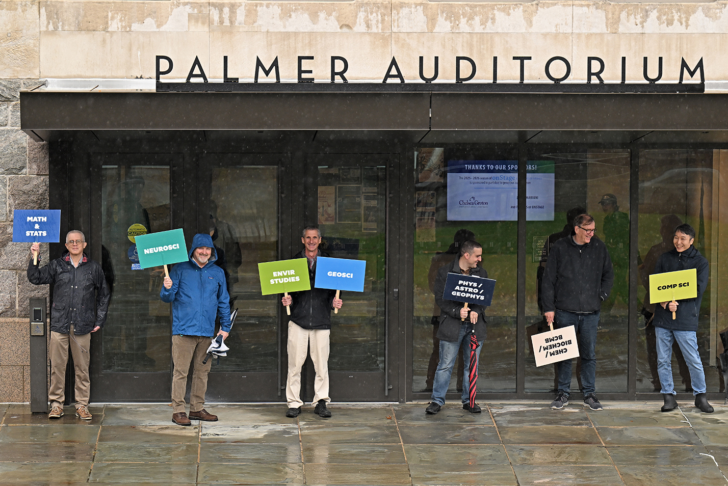 A row of college professors stand holding small signs indicating their academic areas for tours.