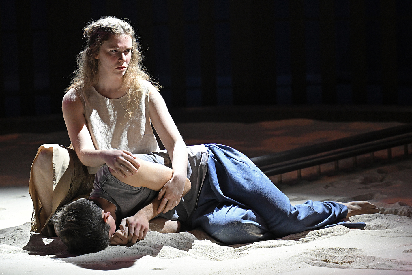 Two student actors, one sitting, one laying prone, perform on a stage covered in sand in a black box theater.
