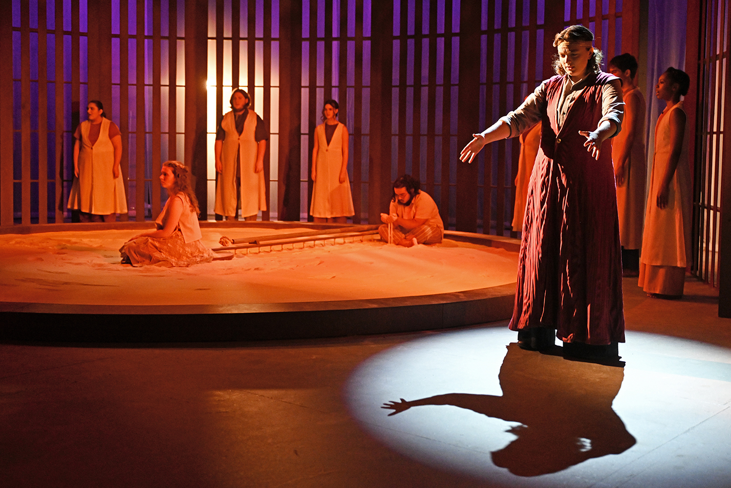A student actor in a royal robe costume stands in a spotlight surrounded by other cast members on stage in black box theater.