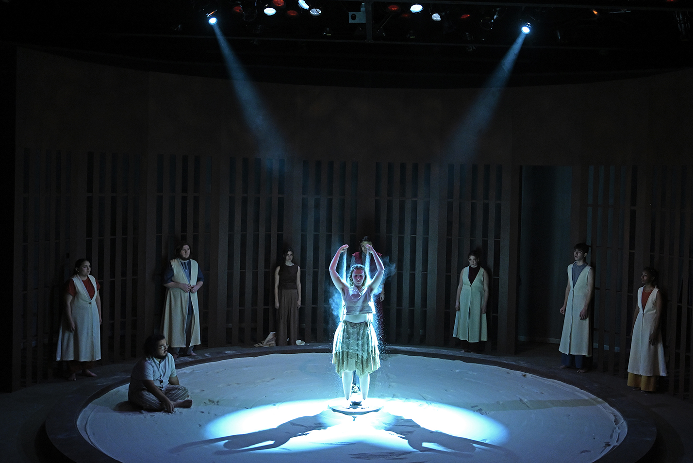 A student actor stands spotlit in the middle of a black box theater stage pouring handfuls of sand over her shoulders as the remaining cast watches from the shadows.