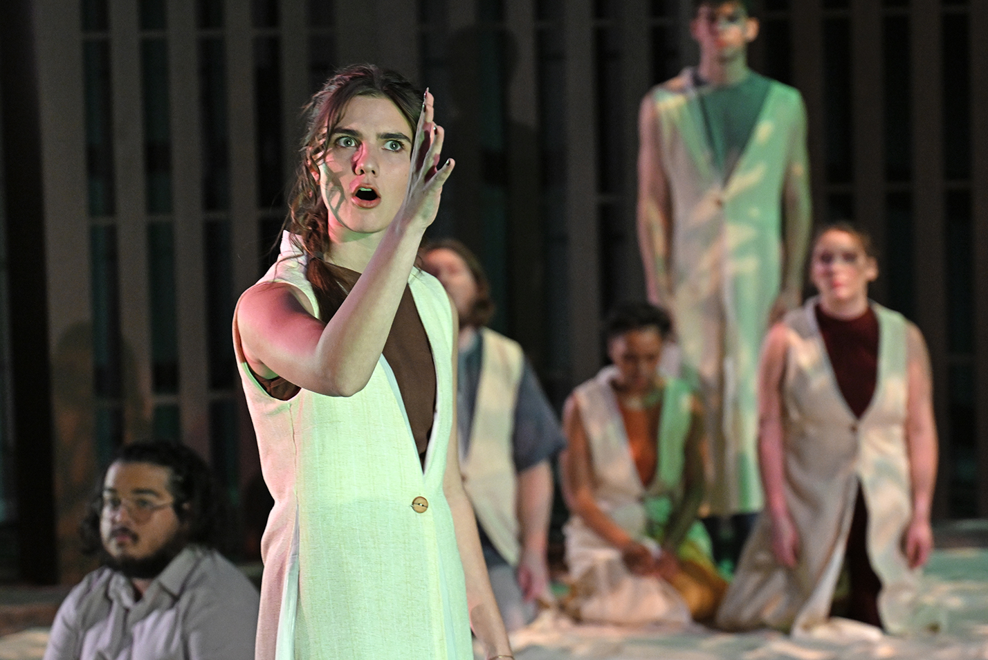 A student actor in a white robe gestures with her right hand up toward the sky as other actors look on in the background.
