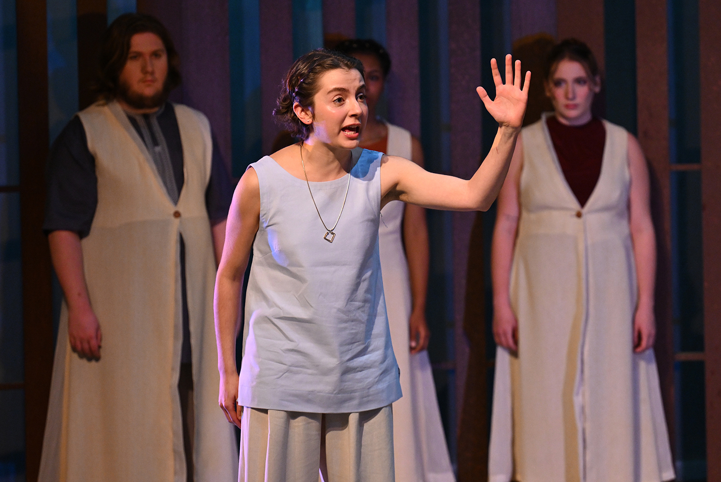 A student actor in a light blue tunic gestures with her left hand as other actors look on in the background.