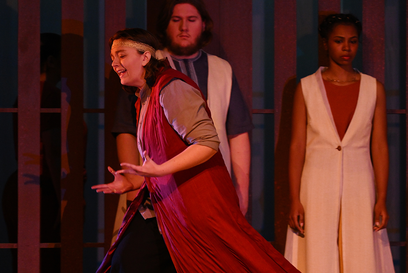 A student actor in a royal robe costume gestures and strides by other cast members on stage in black box theater.