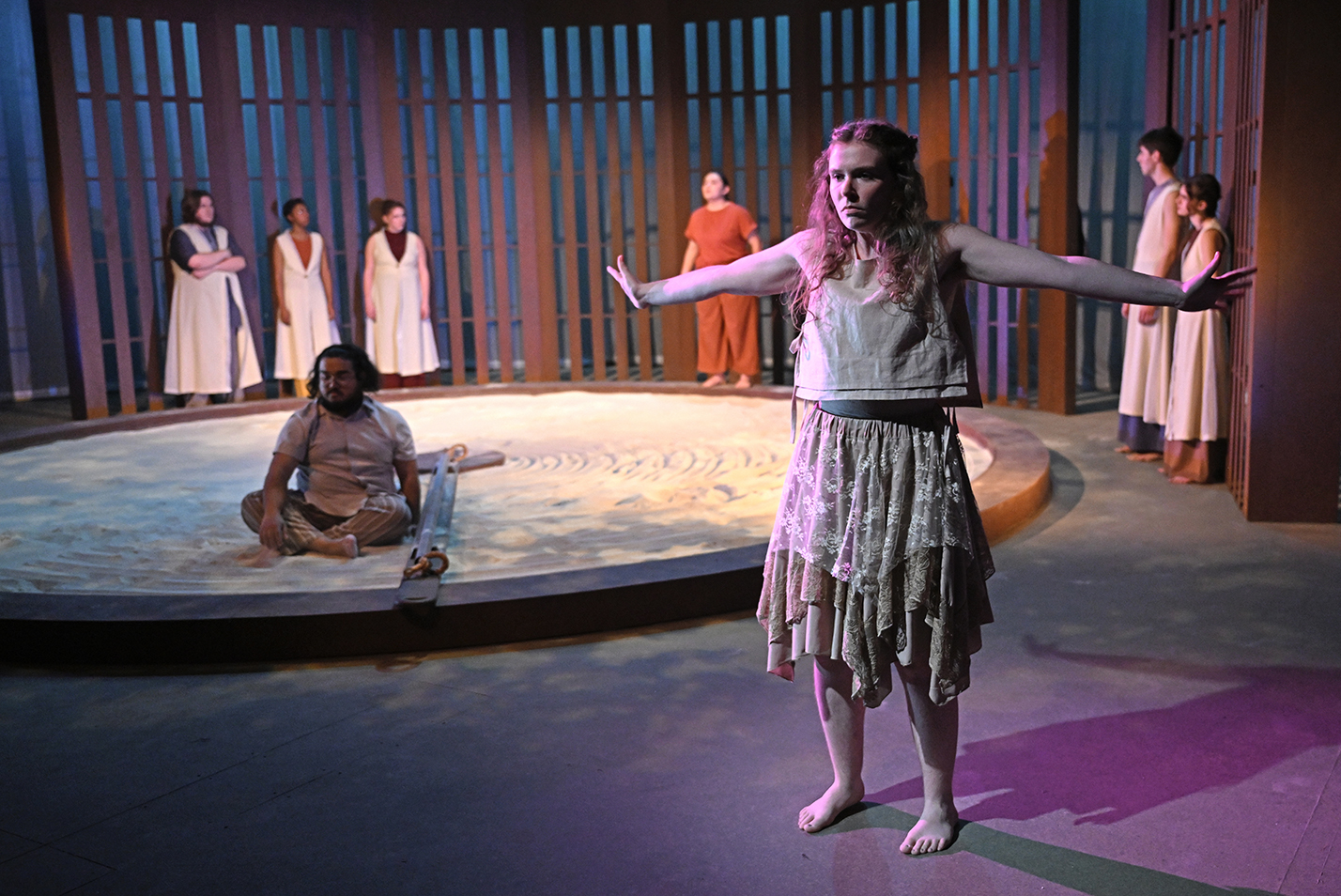 A student actor stands downstage with her arms out in front of the remaining cast on stage in a black box theater.