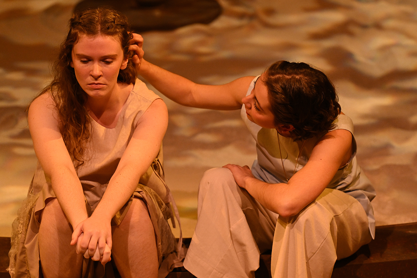 One student actor gently caresses another's hair as they sit on the edge of a stage in a black box theater.