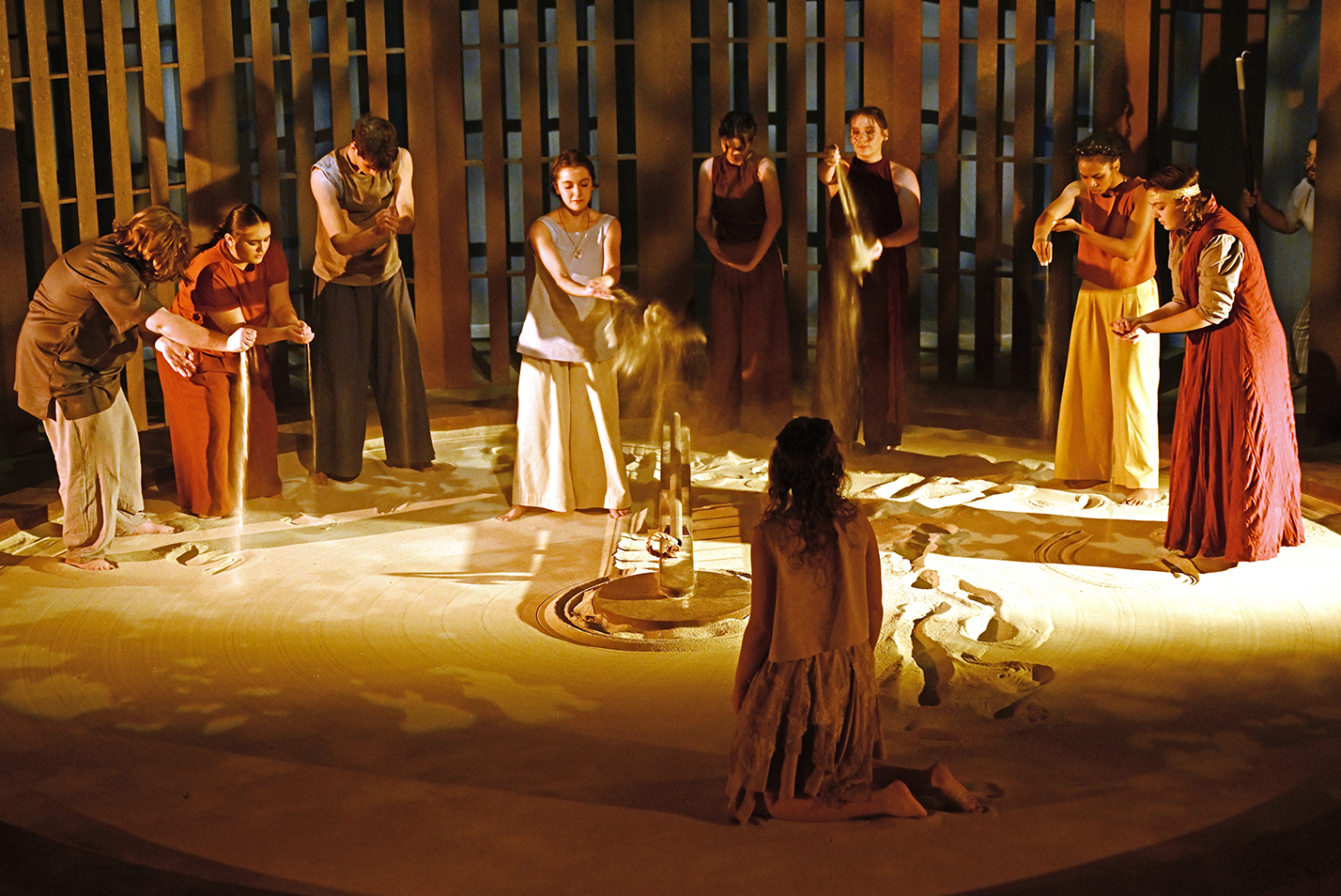 The cast of a play pour sand from their hands on a stage covered in sand for a scene in a black box theater.