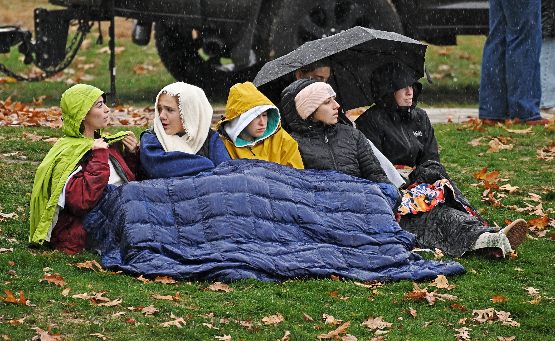 Five female college students sit on a grassy hillside under a blanket and with an umbrella and wearing coats and hoods watching a soccer match in the rain.