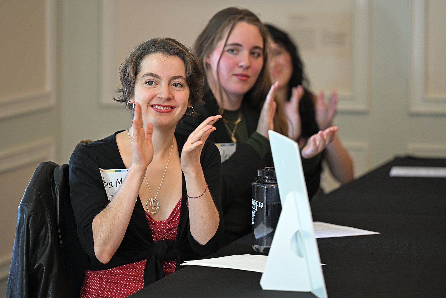 Three college students seated at a table applaud a classmates presentation.
