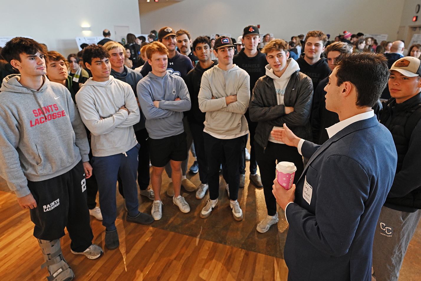 A group of male college students in casual wear listen to classmate dressed in a suit makes a presentation in a large ballroom space.