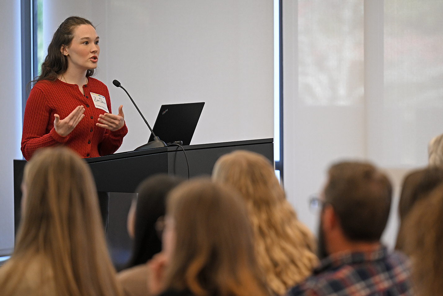 A female student in a red sweater makes a presentation standing at a podium in front of an audience.