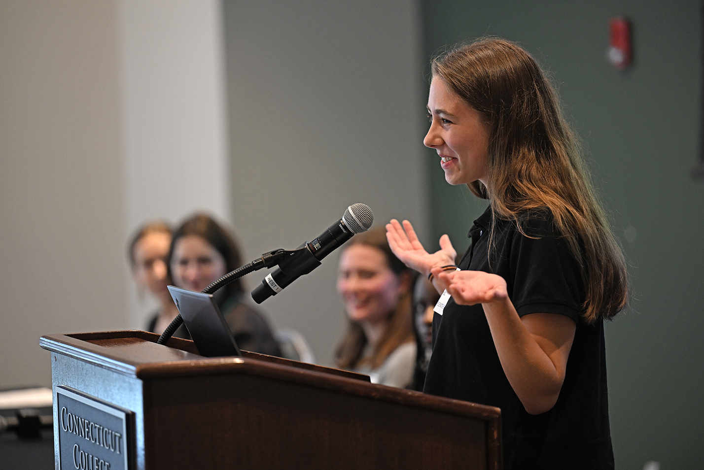 A female student shrugs her shoulders while making a presentation standing at a podium in front of an audience.