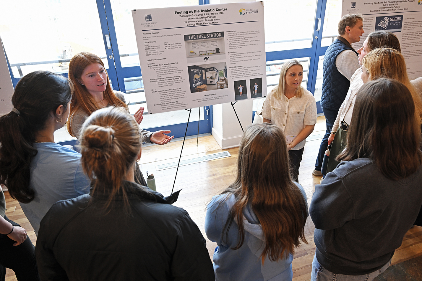 two female college students make a presentation to a group gathered around them in a large ballroom space.