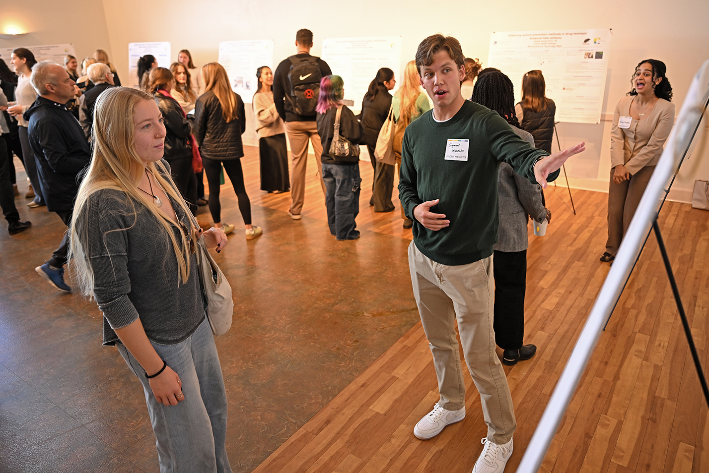 A male college student makes a presentation standing in front of a poster on a tripod stand in a ballroom space.