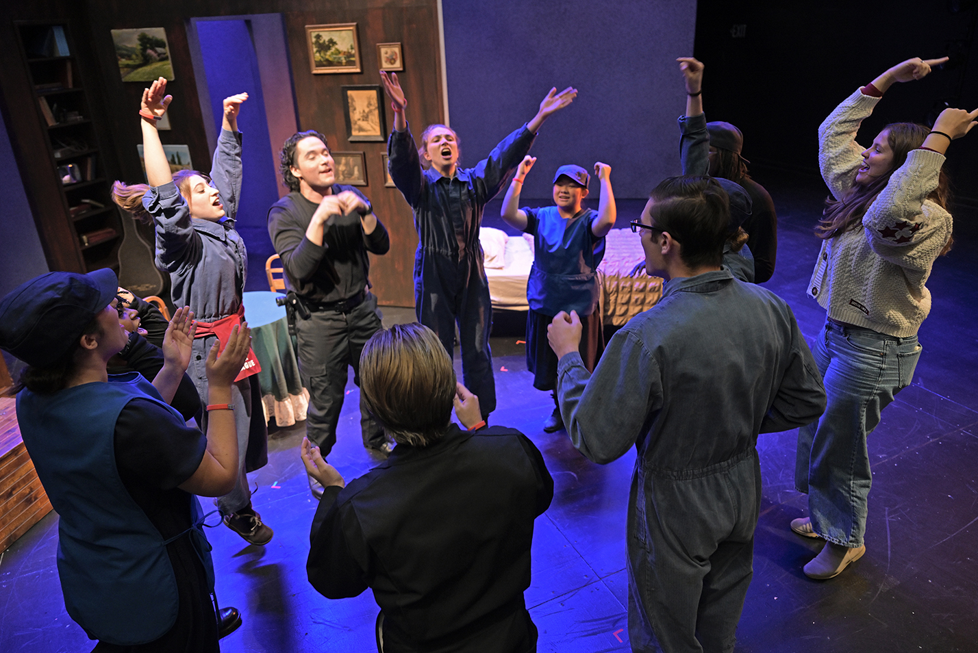 A group of actors in a circle throw their arms into the air and shout as part of a pre-performance warm-up on a darkened stage in a black box theater.