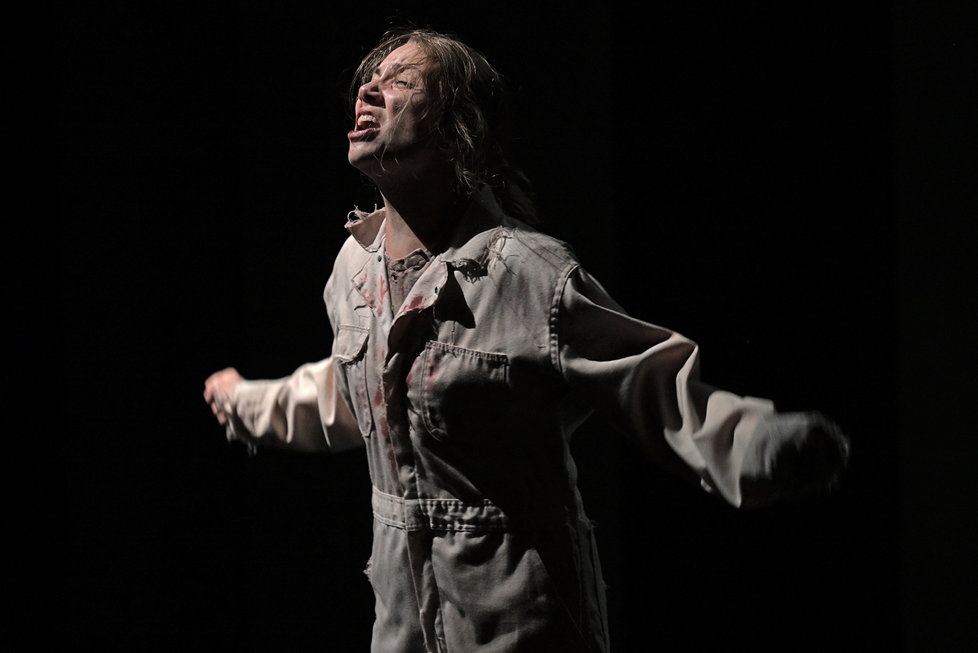 A student actor in a white jumpsuit screams out with arms out on a darkened stage.